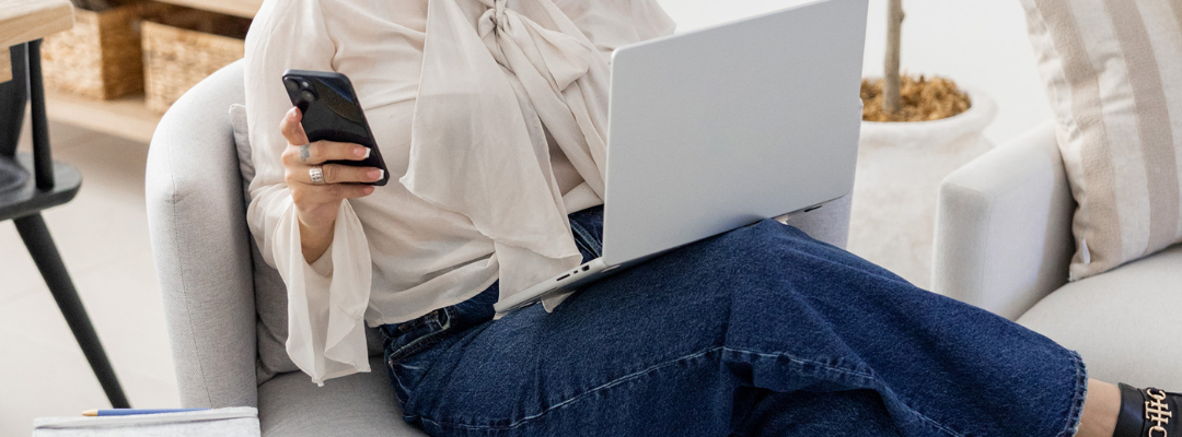 A woman sitting in a sofa holding her laptop on her lap and looking at her phone